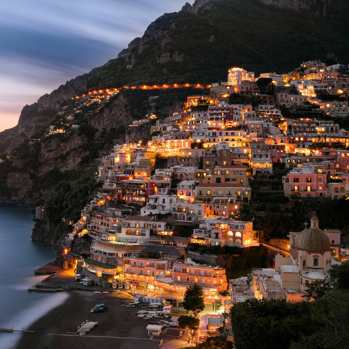 Positano at dusk on the Amalfi coast, the lit hillside town above a calm sea, the place behind LUMIRA Paradiso del Sole