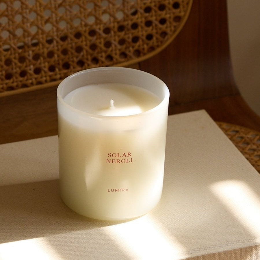 The LUMIRA Solar Neroli candle in its pale vessel against a cane chair