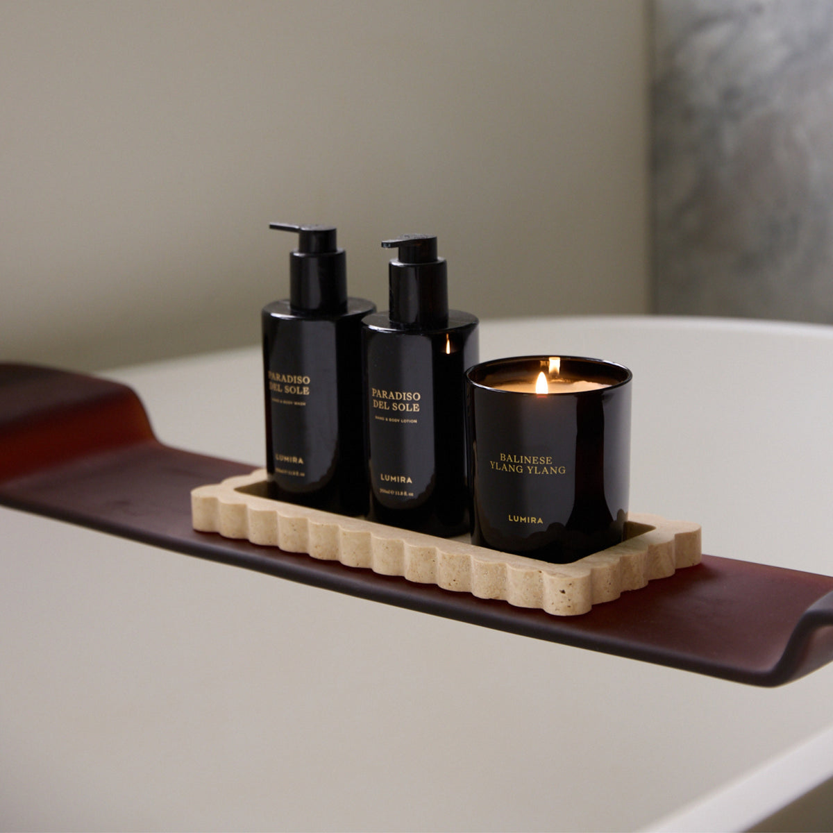 The LUMIRA Balinese Ylang Ylang candle lit on a bath tray beside a hand and body wash and lotion