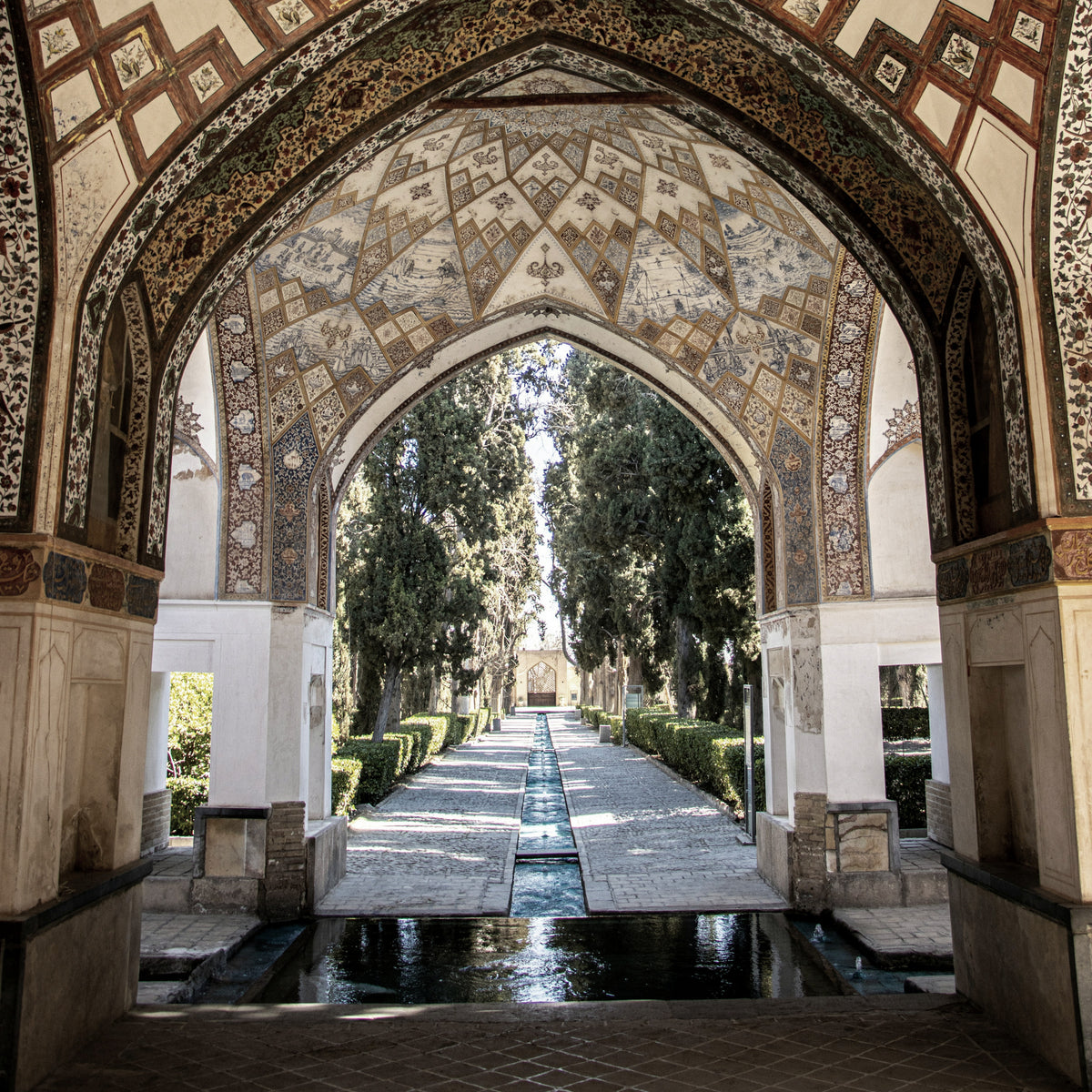A Persian garden framed by an ornate tiled arch, a water channel and cypresses beyond, the place behind LUMIRA Persian Rose
